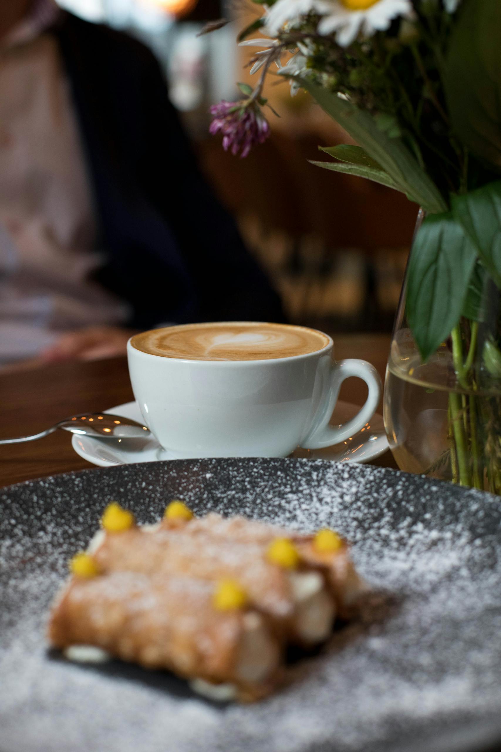 Cup of fresh latte placed near plate with cannoli pastry and vase with flowers during breakfast in cafe